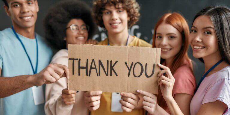 Group of diverse young volunteers smiling at camera, holding card with Thank you lettering while standing in charitable organization office